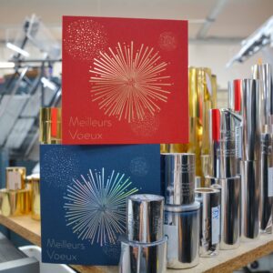 Deux cartes de vœux avec un motif de feu d'artifice en dorure, l'une rouge et l'autre bleu nuit, posées sur un présentoir au milieu de rouleaux métalliques de dorure, dans un atelier d'imprimerie.
