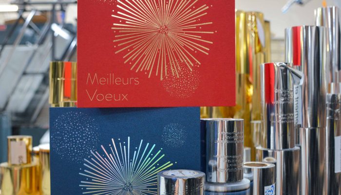 Deux cartes de vœux avec un motif de feu d'artifice en dorure, l'une rouge et l'autre bleu nuit, posées sur un présentoir au milieu de rouleaux métalliques de dorure, dans un atelier d'imprimerie.
