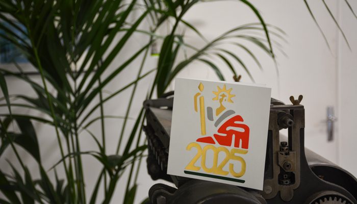 Carte de vœux 2025 avec une illustration stylisée en dorure à chaud or et rouge, représentant une Marianne et un notaire, accompagnée du chiffre '2025' en dorure. La carte est posée sur une ancienne presse d'imprimerie, avec un fond composé de grandes plantes vertes.